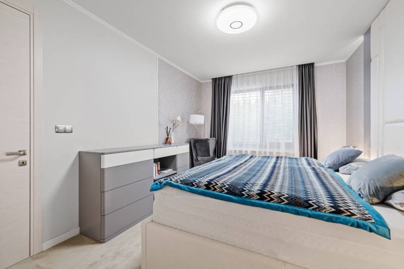 Spálňa v rodinnom dome s posteľou, komodou a veľkým oknom s tmavými záclonami.